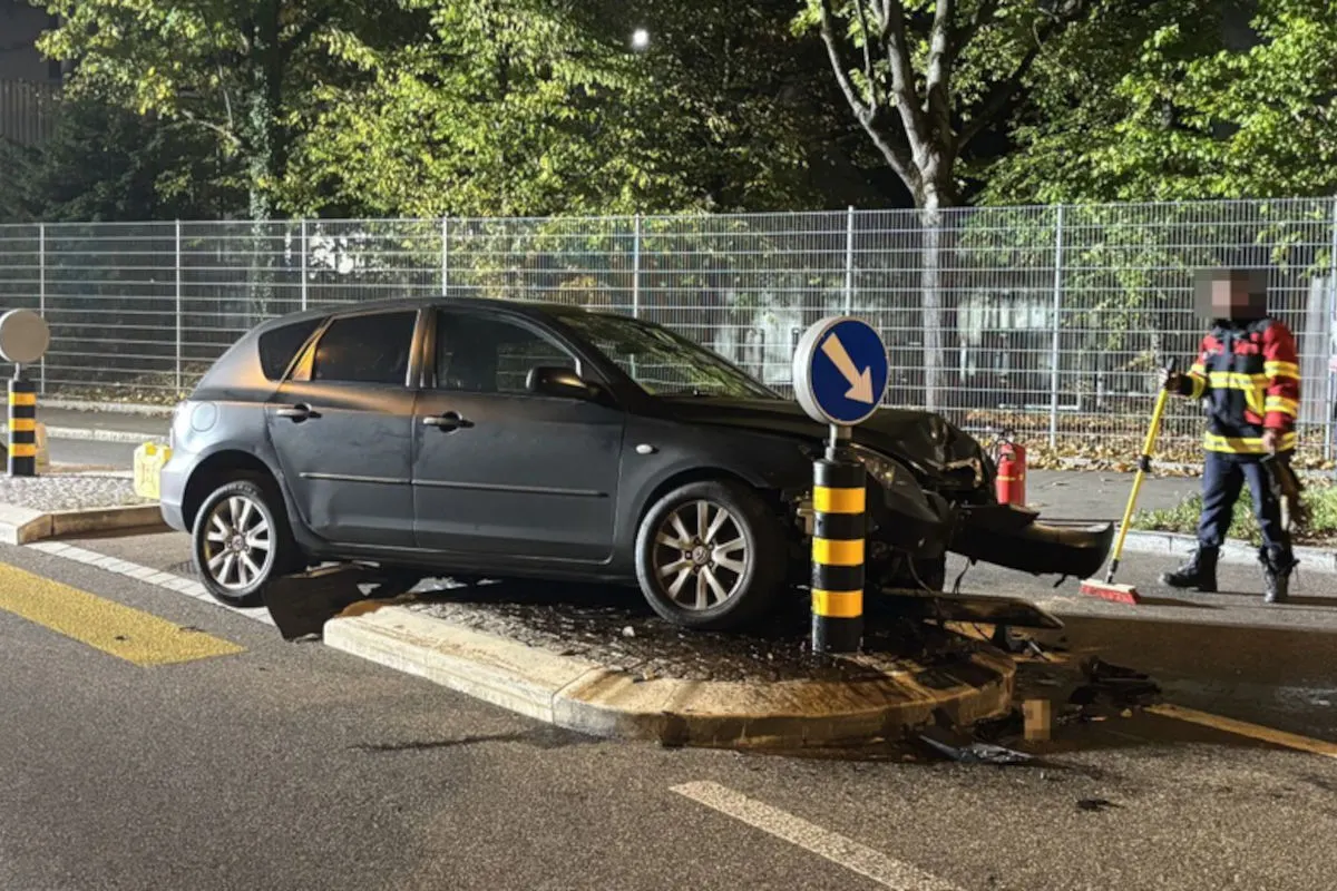Ein dunkles Auto ist auf eine Verkehrsinsel gefahren und hat ein Verkehrsschild gerammt. Eine Person in Warnkleidung ist daneben zu sehen.