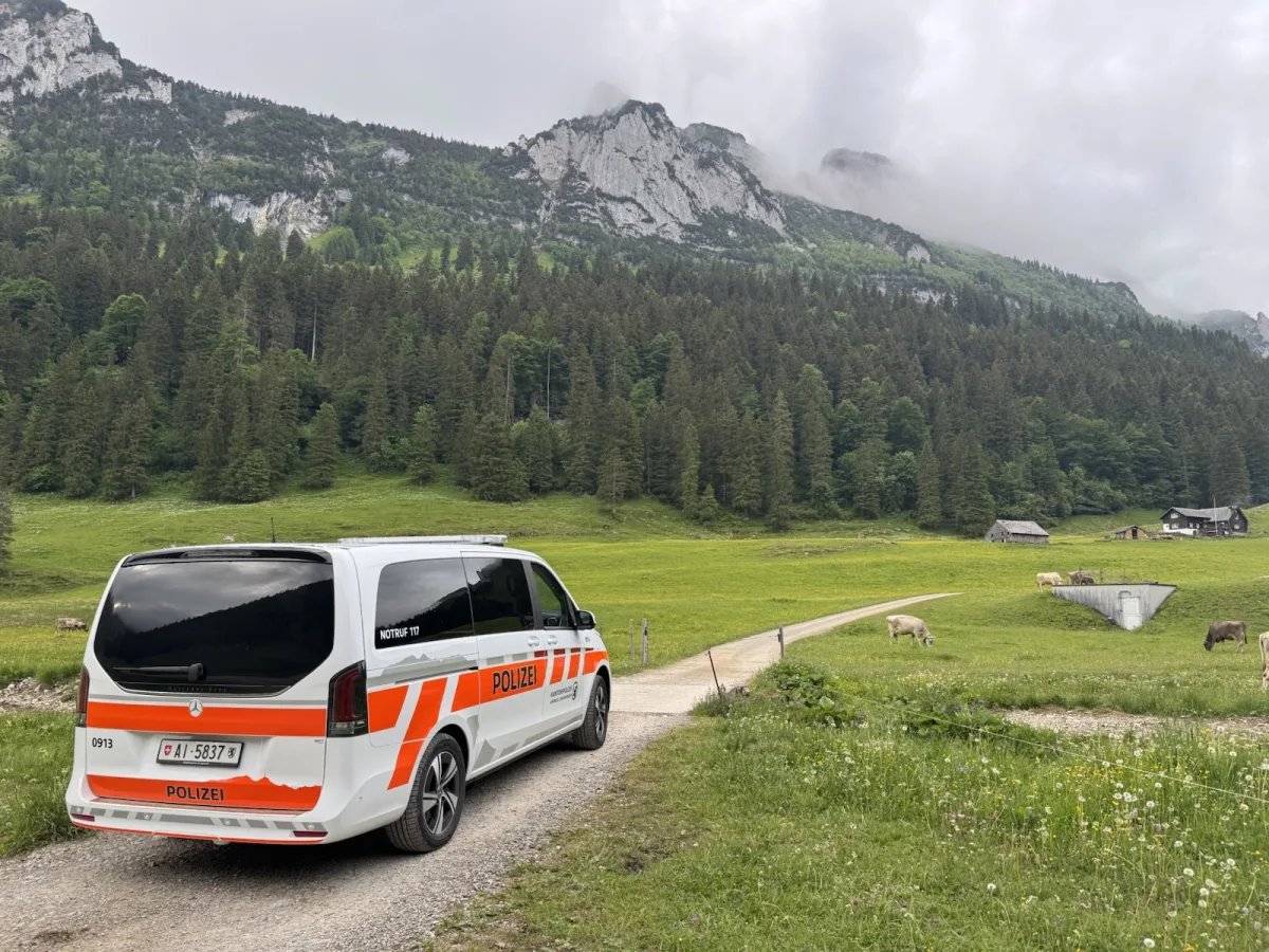 Ein weißer Polizei-Van mit orangen Streifen steht auf einem Schotterweg in einer grünen, alpinen Landschaft mit Bergen im Hintergrund.