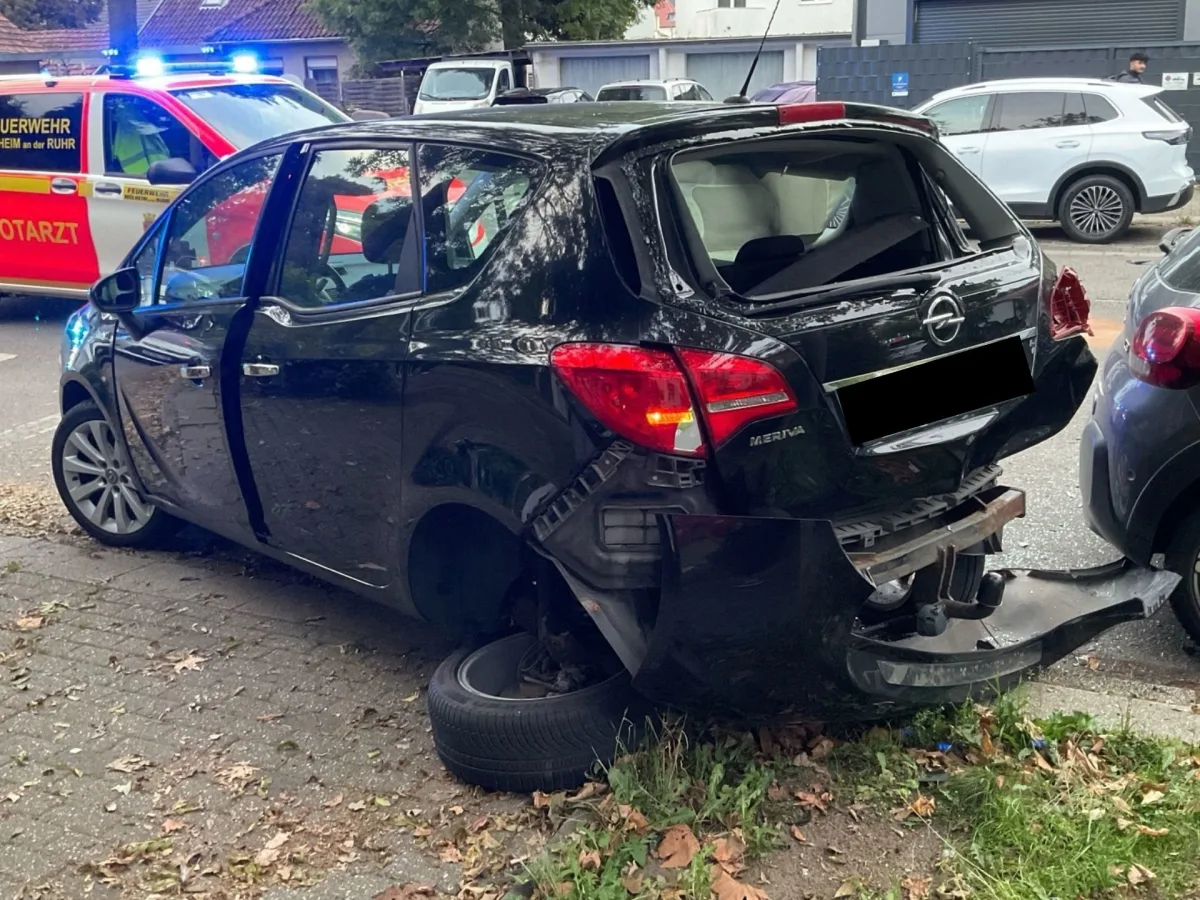 Ein schwarzer Opel Meriva steht auf einem Gehweg mit abgerissener Heckstoßstange. Im Hintergrund ist ein Rettungsdienstfahrzeug mit eingeschaltetem Blaulicht sichtbar.