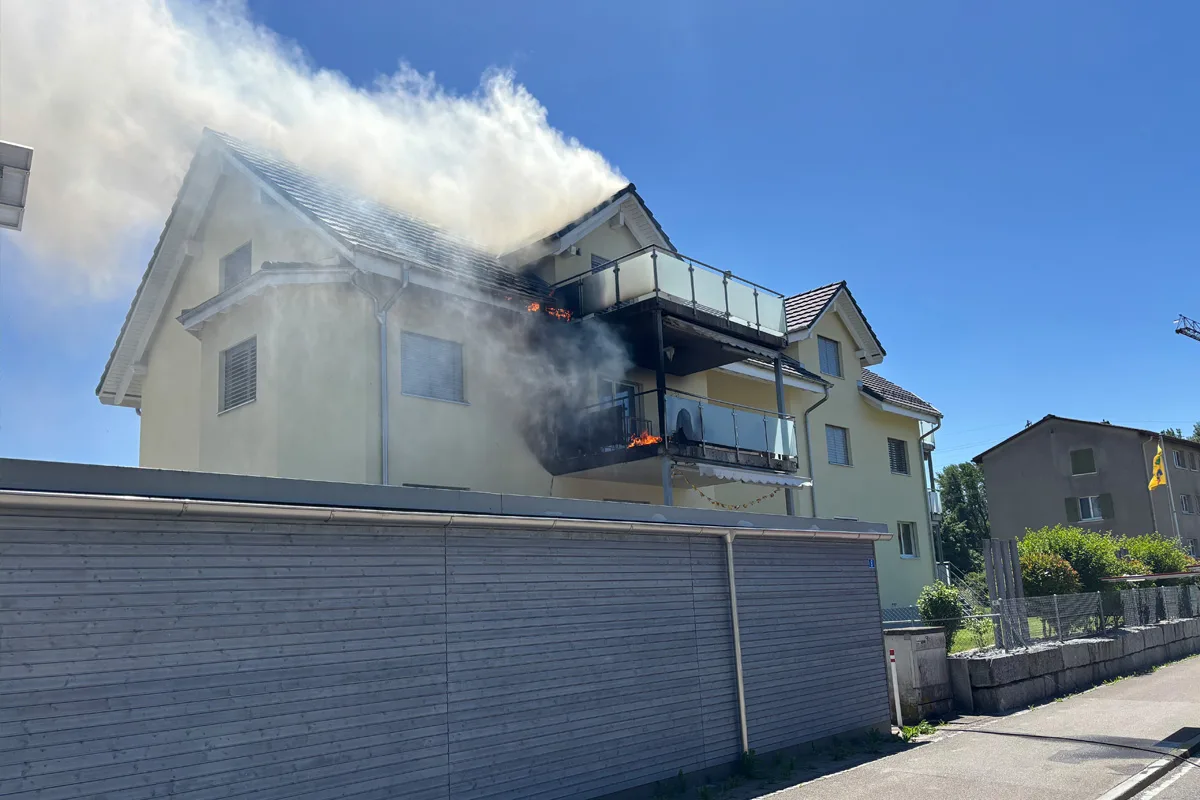 Ein helles Wohnhaus, von dessen Balkon dichter Rauch aufsteigt. Ein kleines Feuer ist auf dem Balkon sichtbar.