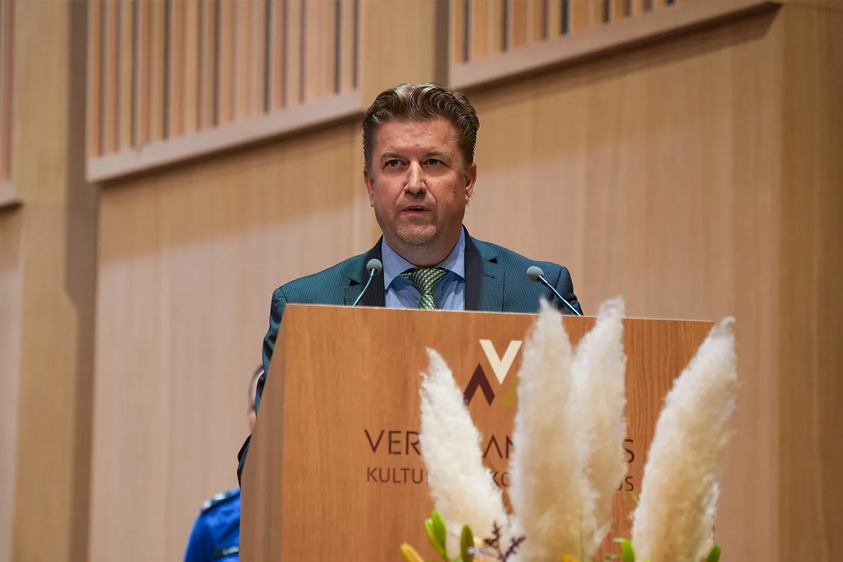 Eine Person hält eine Rede an einem Rednerpult in einem Saal mit Holzverkleidung. Eine weitere Person in blauer Kleidung ist teilweise sichtbar.