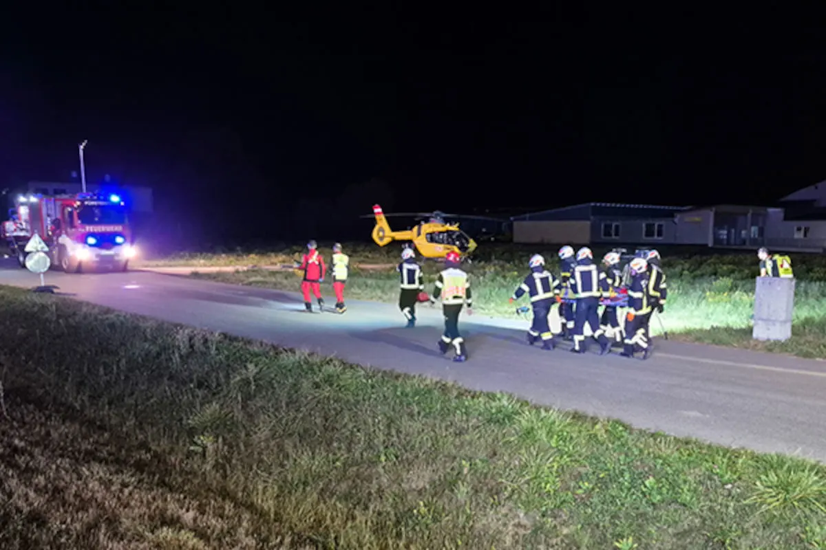 Das Bild zeigt Feuerwehrleute und Rettungsdienste bei Nacht mit einem gelben Hubschrauber im Hintergrund auf einer ländlichen Straße.