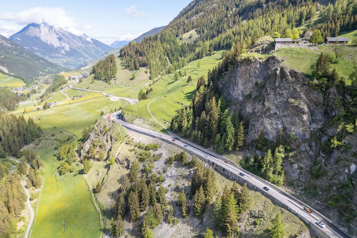 Eine kurvenreiche Straße entlang eines Berghangs, umgeben von Wäldern und Wiesen, ist von mehreren Fahrzeugen befahren.