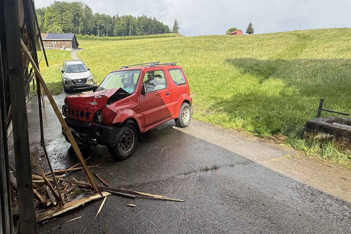 Ein rotes Auto mit stark beschädigter Front steht in einer ländlichen Gegend neben einer eingestürzten Holzkonstruktion. Im Hintergrund ist eine grüne Wiese sichtbar.