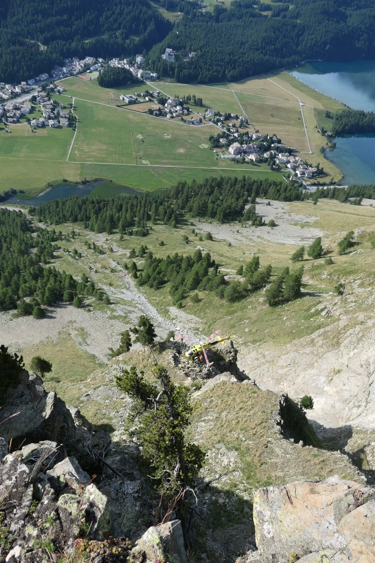 Ein gelber Rettungshubschrauber liegt auf einem felsigen Bergrücken, umgeben von Bäumen, mit einer Ortschaft im Hintergrund.