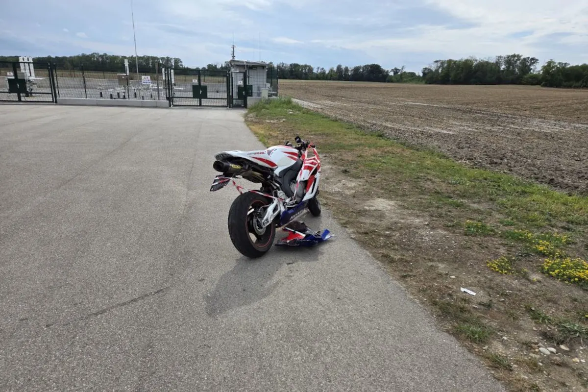 Ein beschädigtes Motorrad mit rot-weißer Lackierung steht auf einer Landstraße. Im Hintergrund sind ein Zaun und Tor zu sehen.
