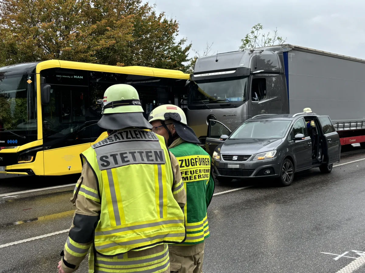 Ein gelber Bus und ein grauer LKW sind bei einem Unfall auf der Straße kollidiert. Feuerwehrleute in Schutzkleidung stehen im Vordergrund.