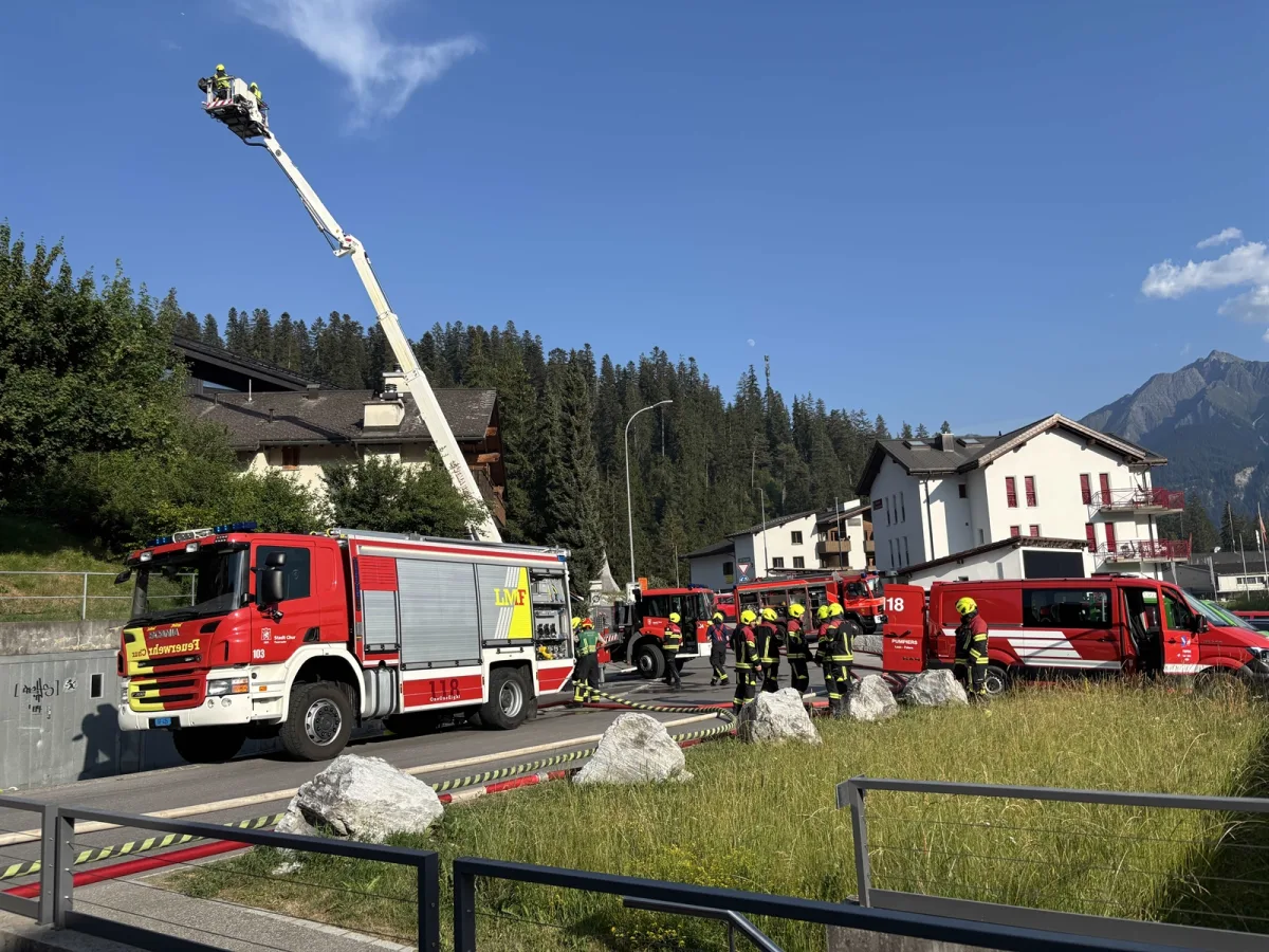 Mehrere Feuerwehrfahrzeuge und Einsatzkräfte in einem ländlichen Gebiet vor einem Haus mit Berge im Hintergrund. Eine Leiter ist ausgefahren.