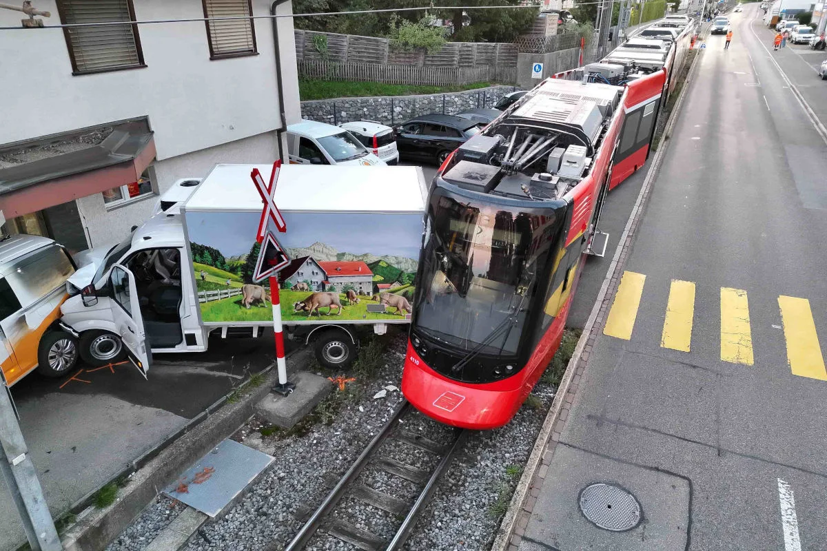 Ein roter Straßenbahnwagen ist an einem Bahnübergang mit einem Lieferwagen kollidiert, das Tor ist beschädigt. Der Lieferwagen hat eine ländliche Szene auf der Seite abgebildet und stößt gegen ein weiteres, parkendes Fahrzeug.