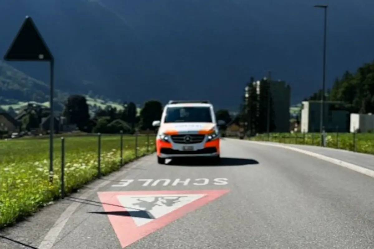Ein Rettungswagen fährt auf einer kurvigen Landstraße in einer grünen, ländlichen Umgebung. Auf der Straße ist eine rote Markierung mit dem Wort 'SCHULE' und einem Warnsymbol zu sehen.