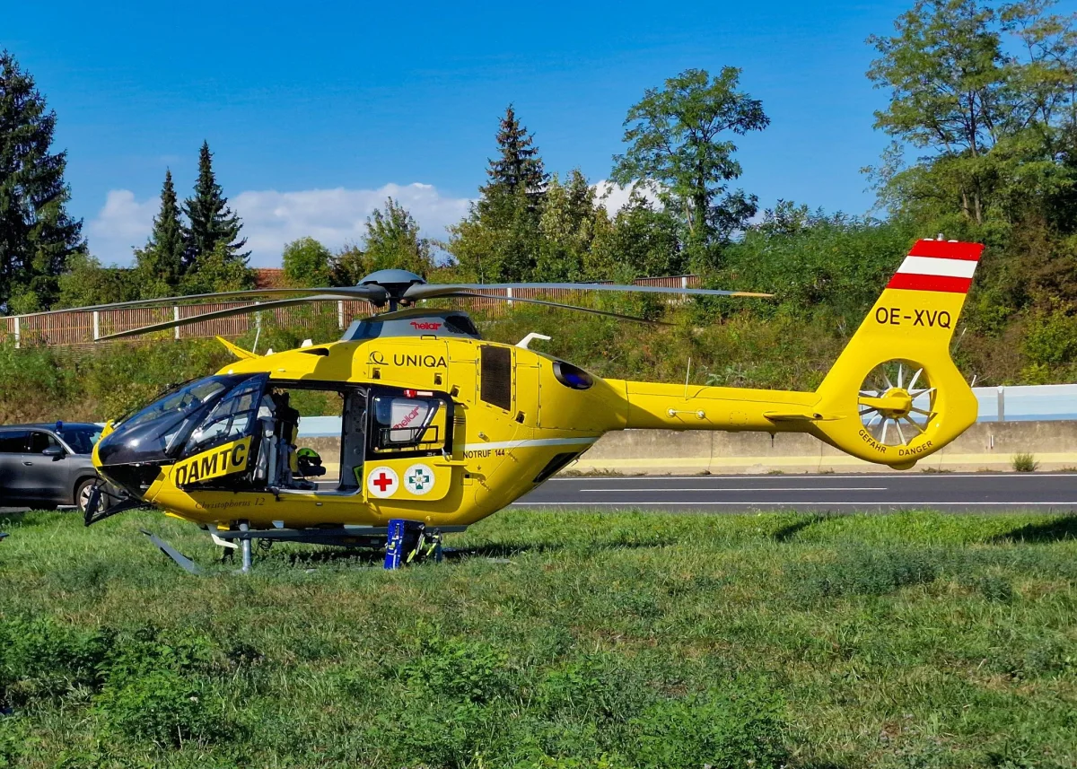 Ein gelber ÖAMTC-Rettungshubschrauber steht auf einer Wiese neben einer Straße. Im Hintergrund sind Bäume und der Himmel zu sehen.