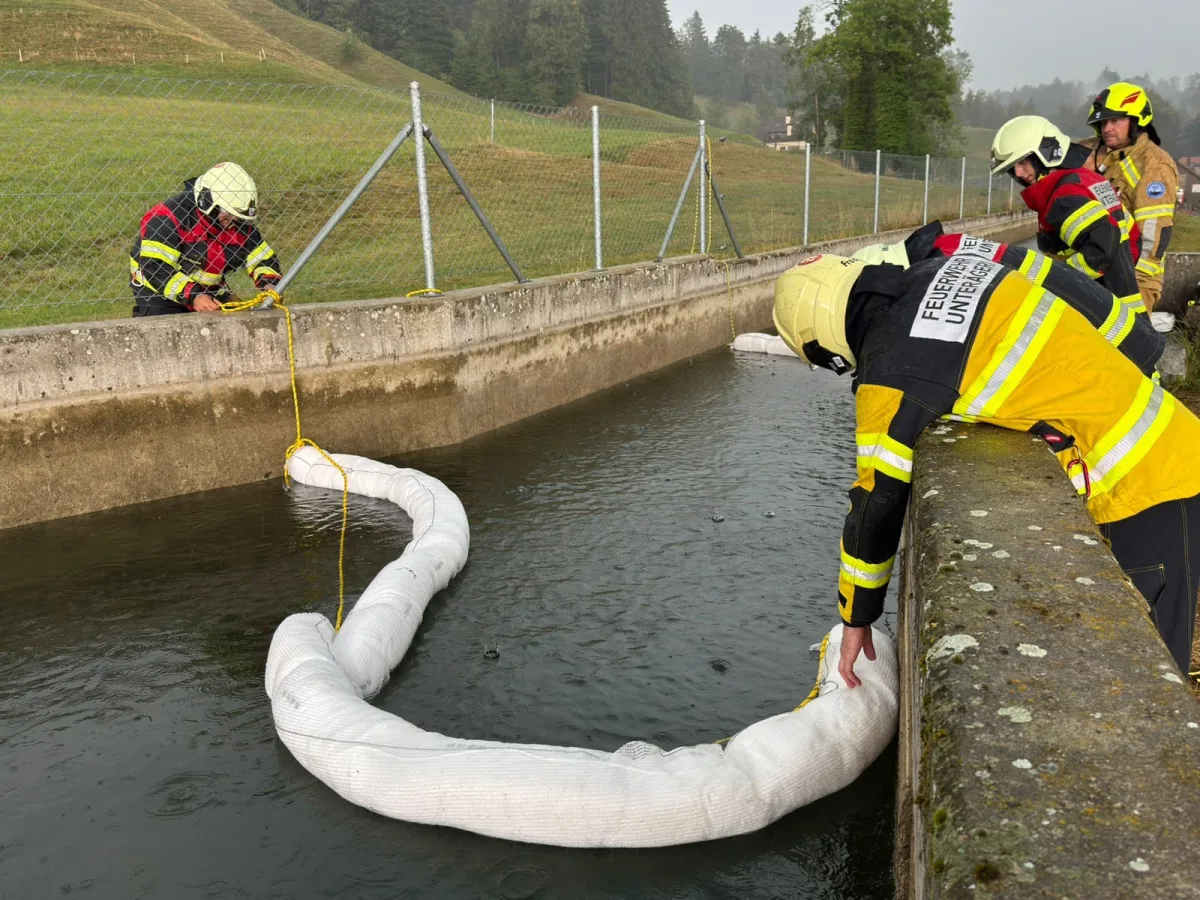Mehrere Feuerwehrleute sichern eine weiße Ölsperre entlang eines Wassergrabens. Sie tragen gelbe und rote Schutzkleidung.