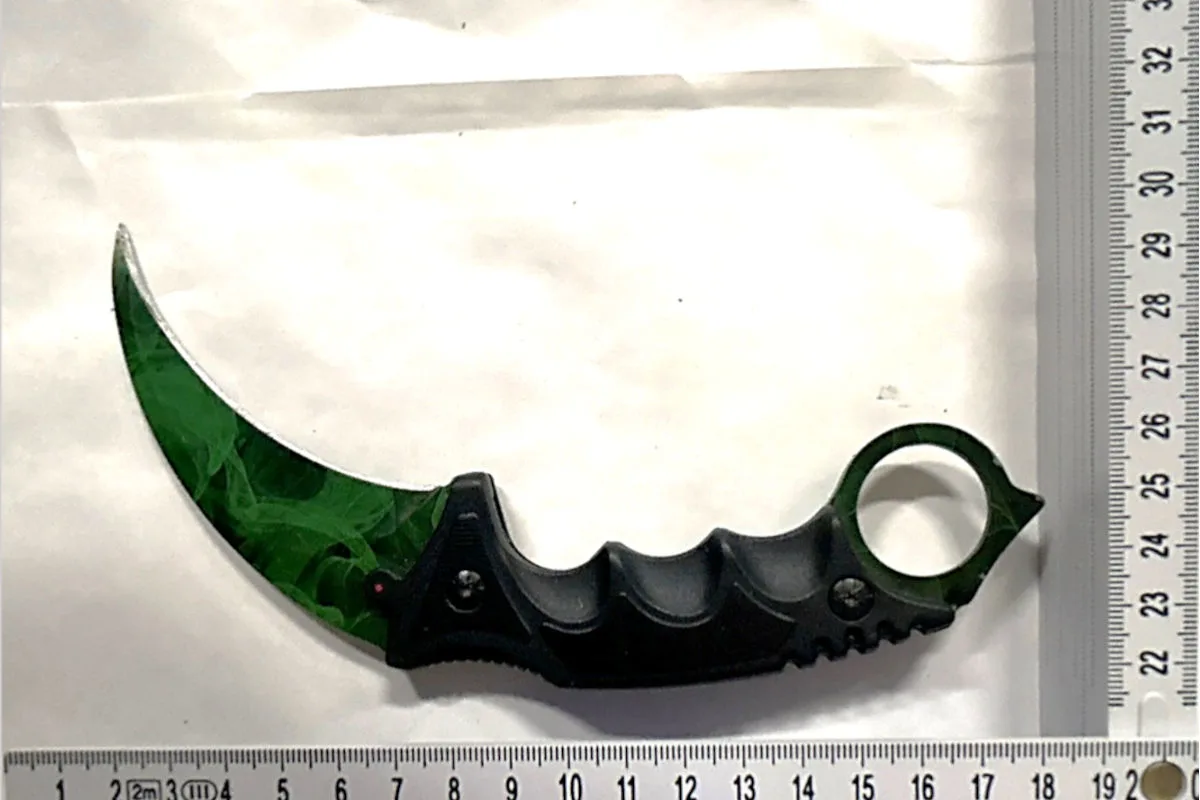 Ein grünes Karambit-Messer mit schwarzem Griff liegt auf einem Lineal zur Größenabschätzung. Das Messer hat eine gebogene Klinge und einen ringförmigen Griff.