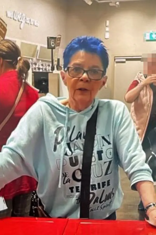Eine Frau mit blauen Haaren und Brille steht an einem Ladentisch, während andere Personen im Hintergrund zu sehen sind.