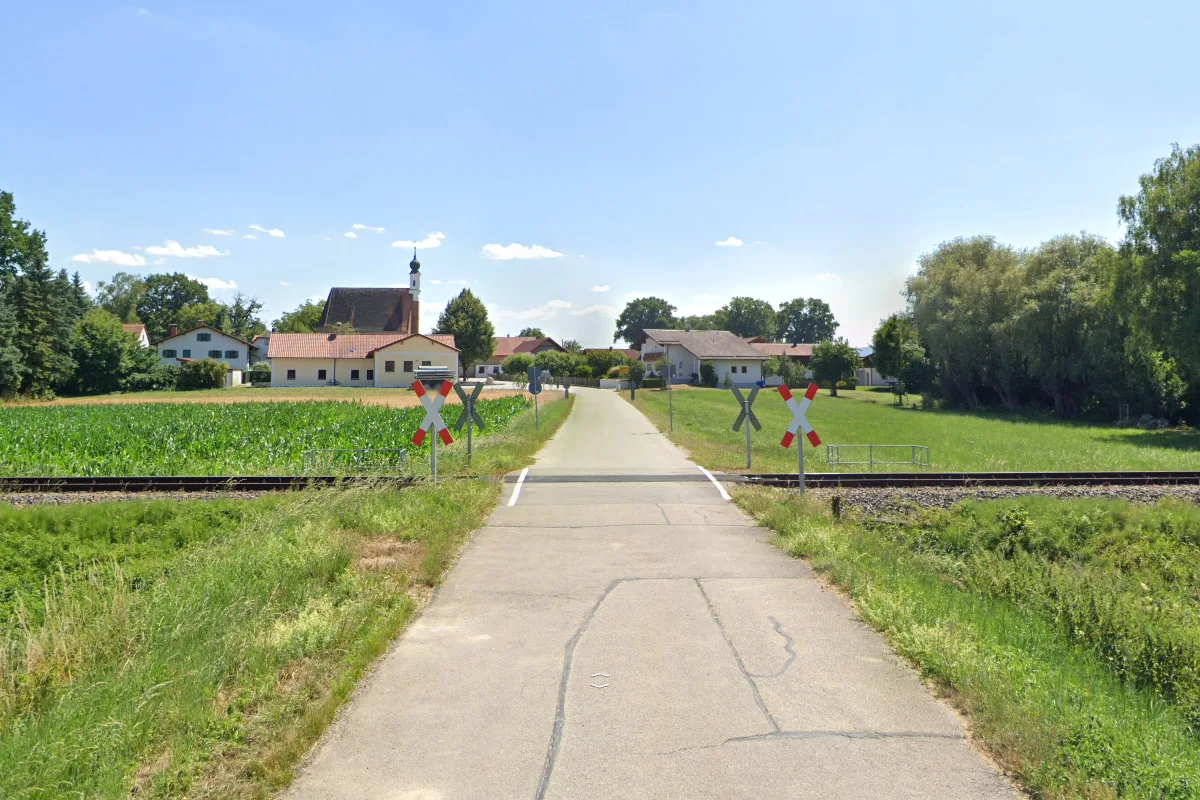 Ein unbeschrankter Bahnübergang führt über eine einzelne Bahngleisstrecke. Im Hintergrund sind mehrere Häuser und eine Kirche zu sehen.