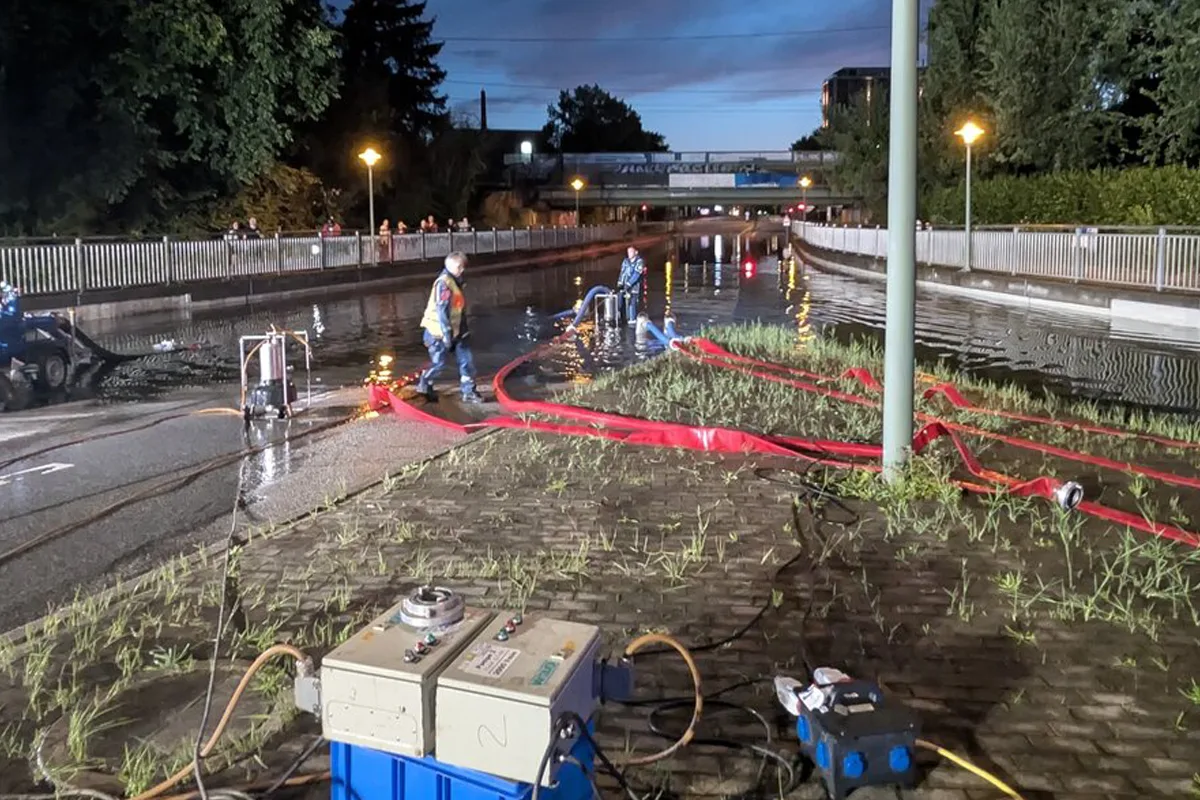 Mehrere Einsatzkräfte stehen auf einer überfluteten Straße bei Nacht und pumpen Wasser ab. Schläuche und Ausrüstung sind sichtbar.
