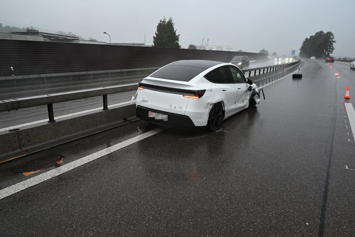 Ein weißes Auto mit sichtbarem Unfallschaden steht auf einer regennassen Autobahn. Verkehrskegel markieren den Unfallbereich.