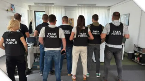 Eine Gruppe von Personen, die ‚Task Force‘-Westen tragen, steht mit dem Rücken zur Kamera in einem Büro.