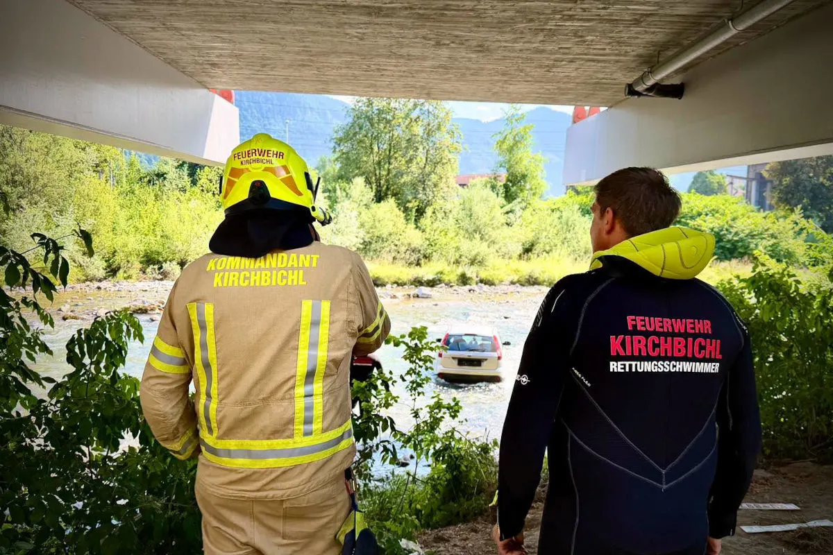Zwei Personen von hinten im Feuerwehreinsatz unter einer Brücke, blicken auf ein Auto, das in einem Fluss steckt.