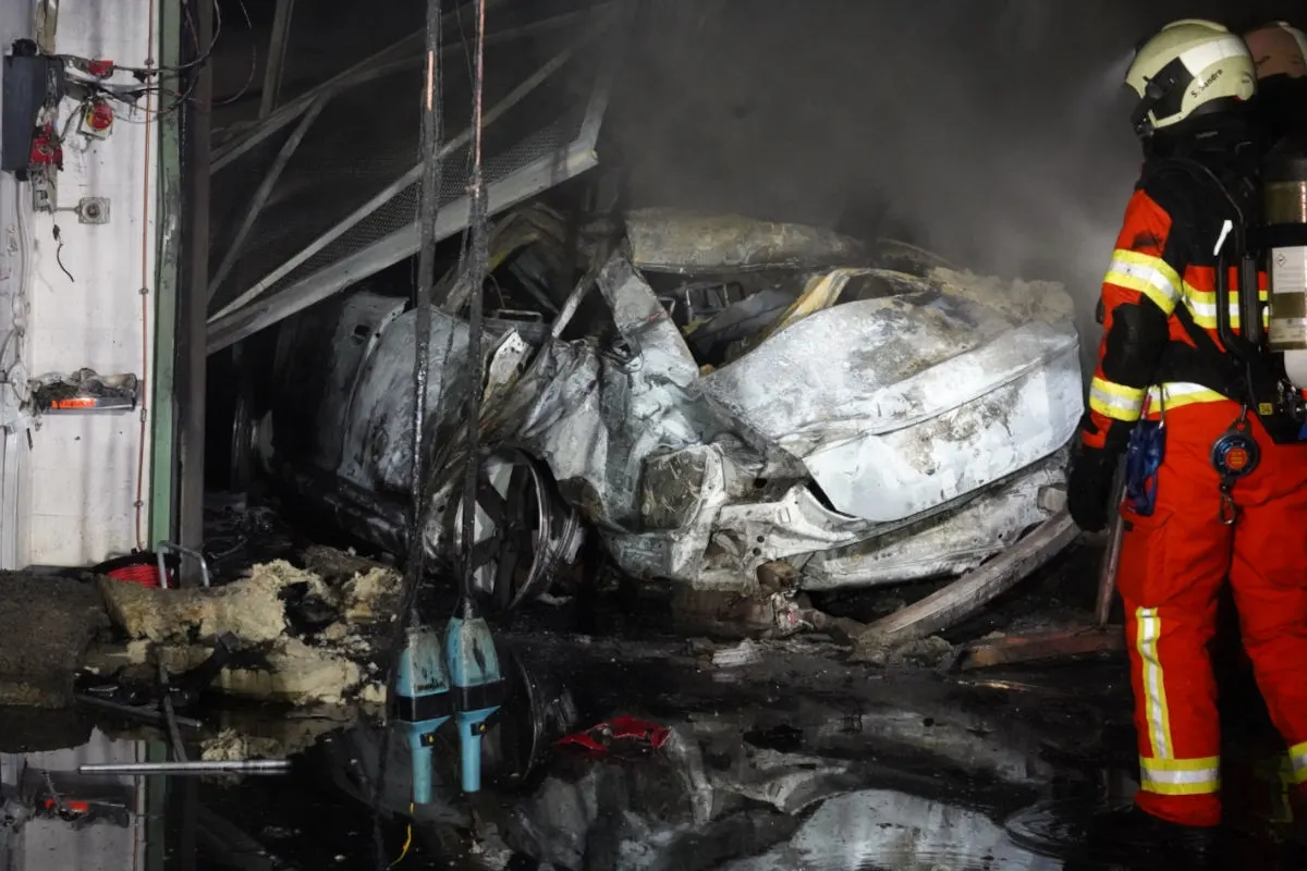 Das Bild zeigt einen Feuerwehrmann in Schutzkleidung vor einem ausgebrannten Autowrack in einem Gebäude. Der Boden ist nass, und das Auto ist stark beschädigt.