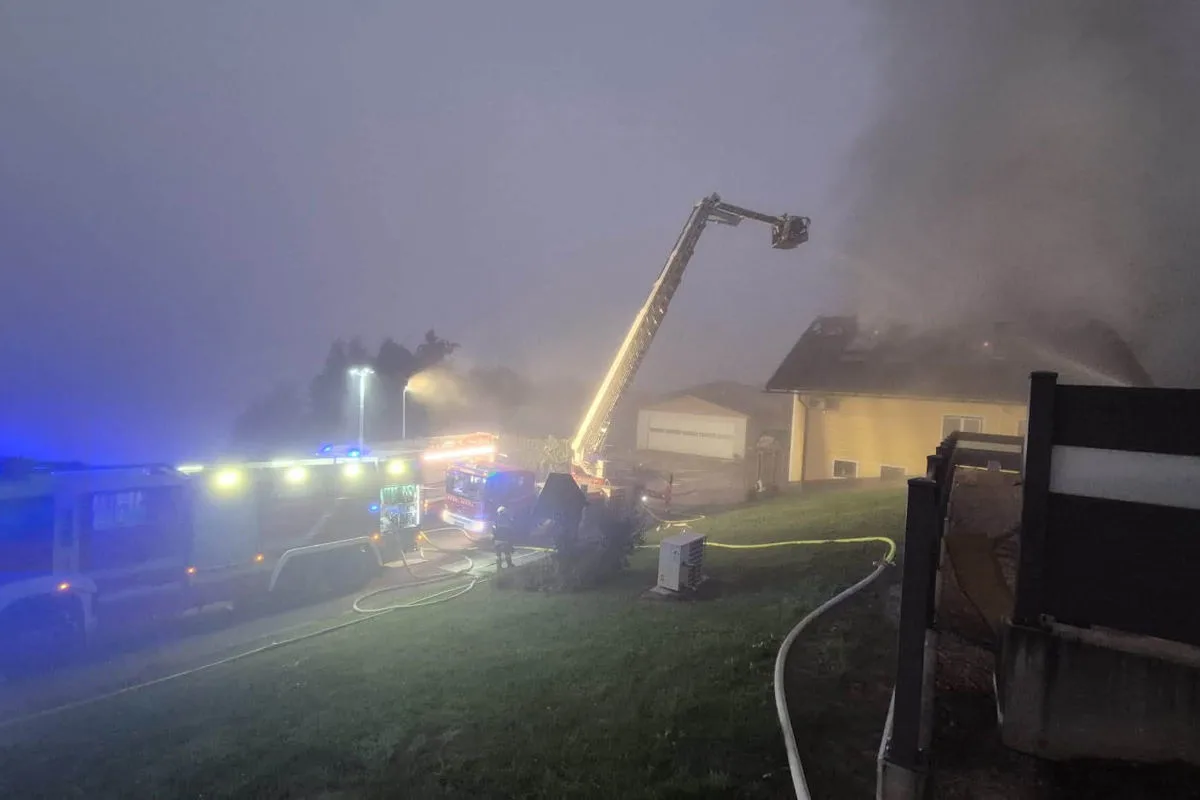 Eine Feuerwehr mit Drehleiter bekämpft einen Brand an einem Wohnhaus. Rauch steigt vom Dach auf, während Schläuche ausgelegt sind.