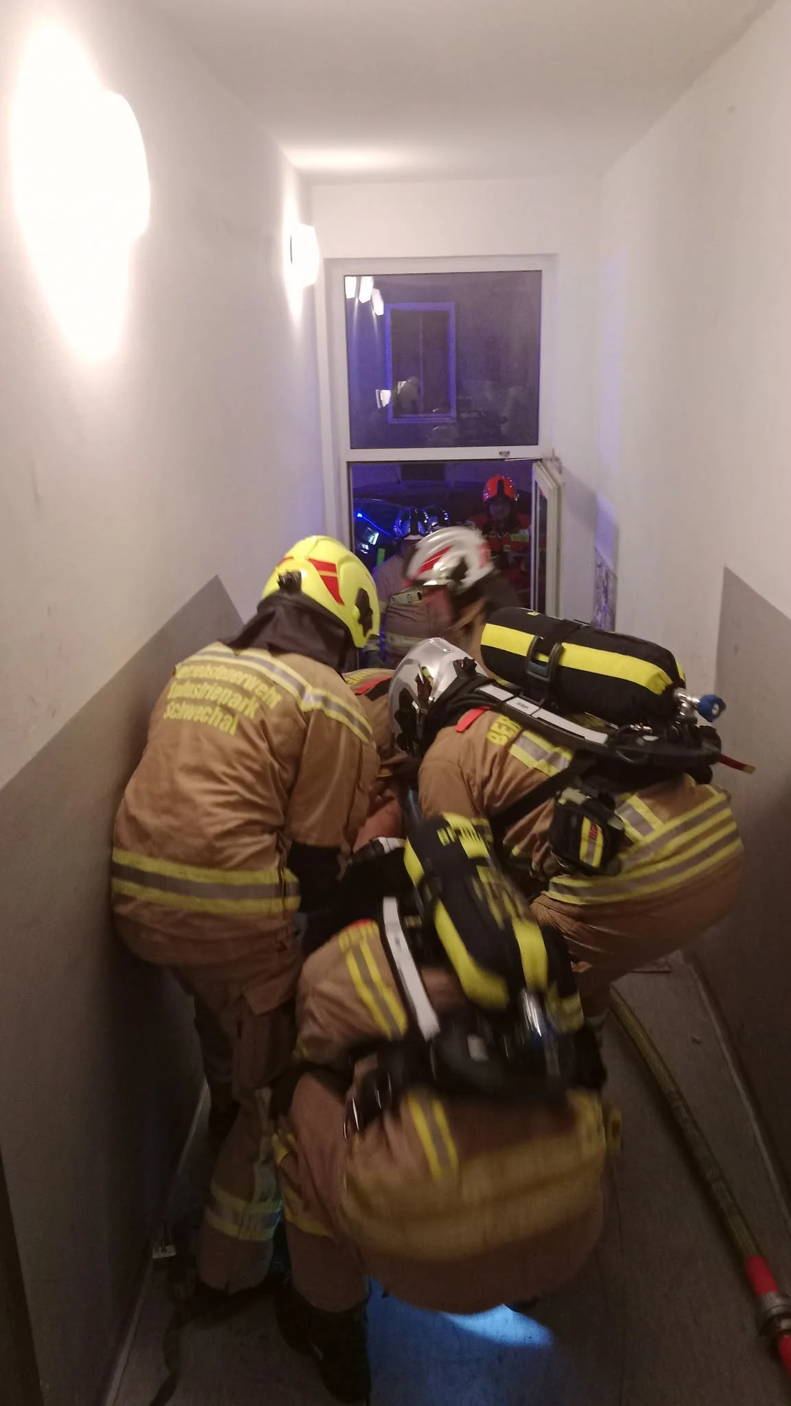 Mehrere Feuerwehrleute in voller Schutzmontur arbeiten in einem engen Treppenhaus. Im Hintergrund sind Blaulichter zu sehen.