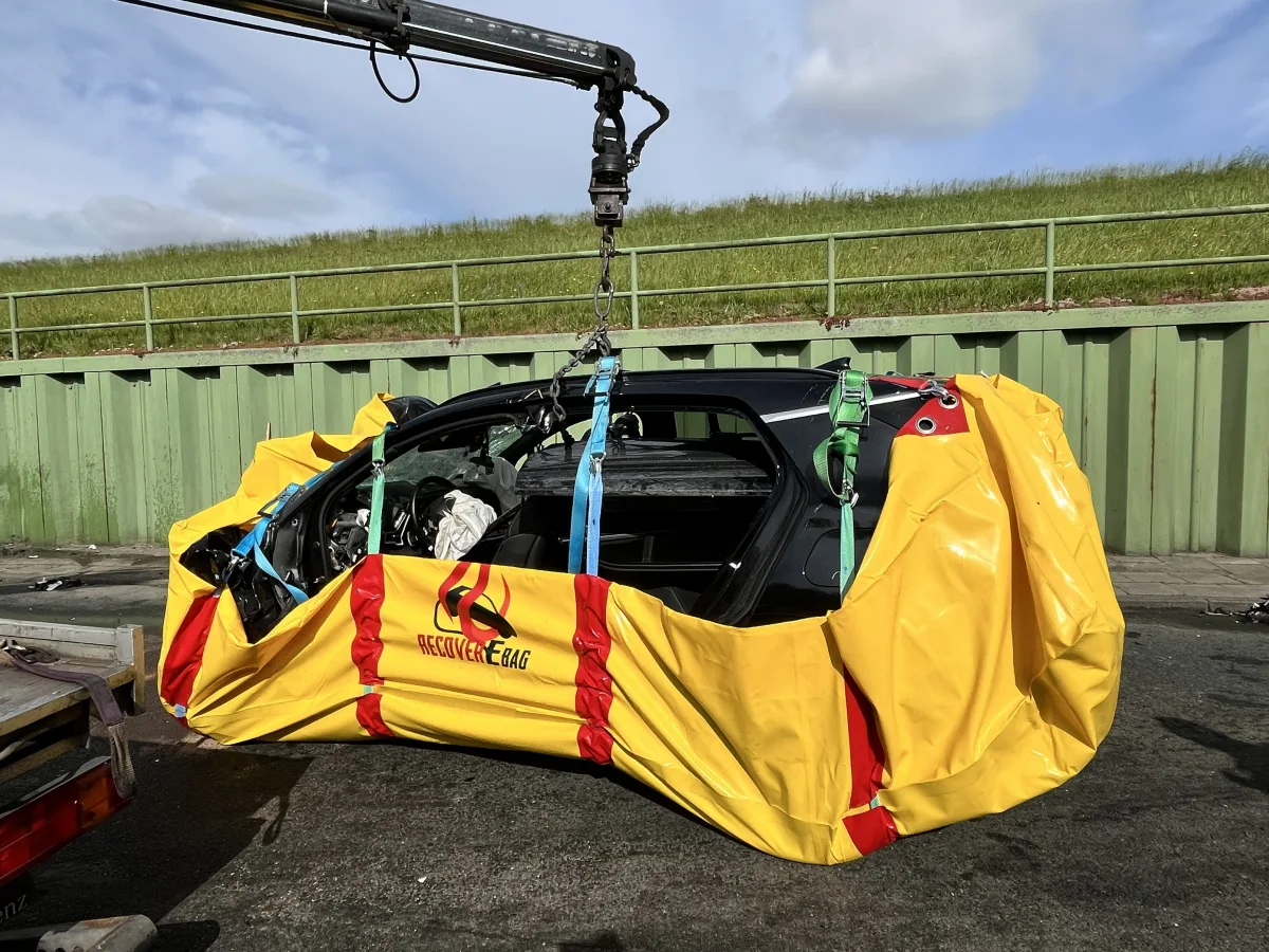 Ein stark beschädigtes Auto wird mithilfe eines Krans und einem gelben Schutzsack angehoben. Im Hintergrund ist eine grüne Wand zu sehen.