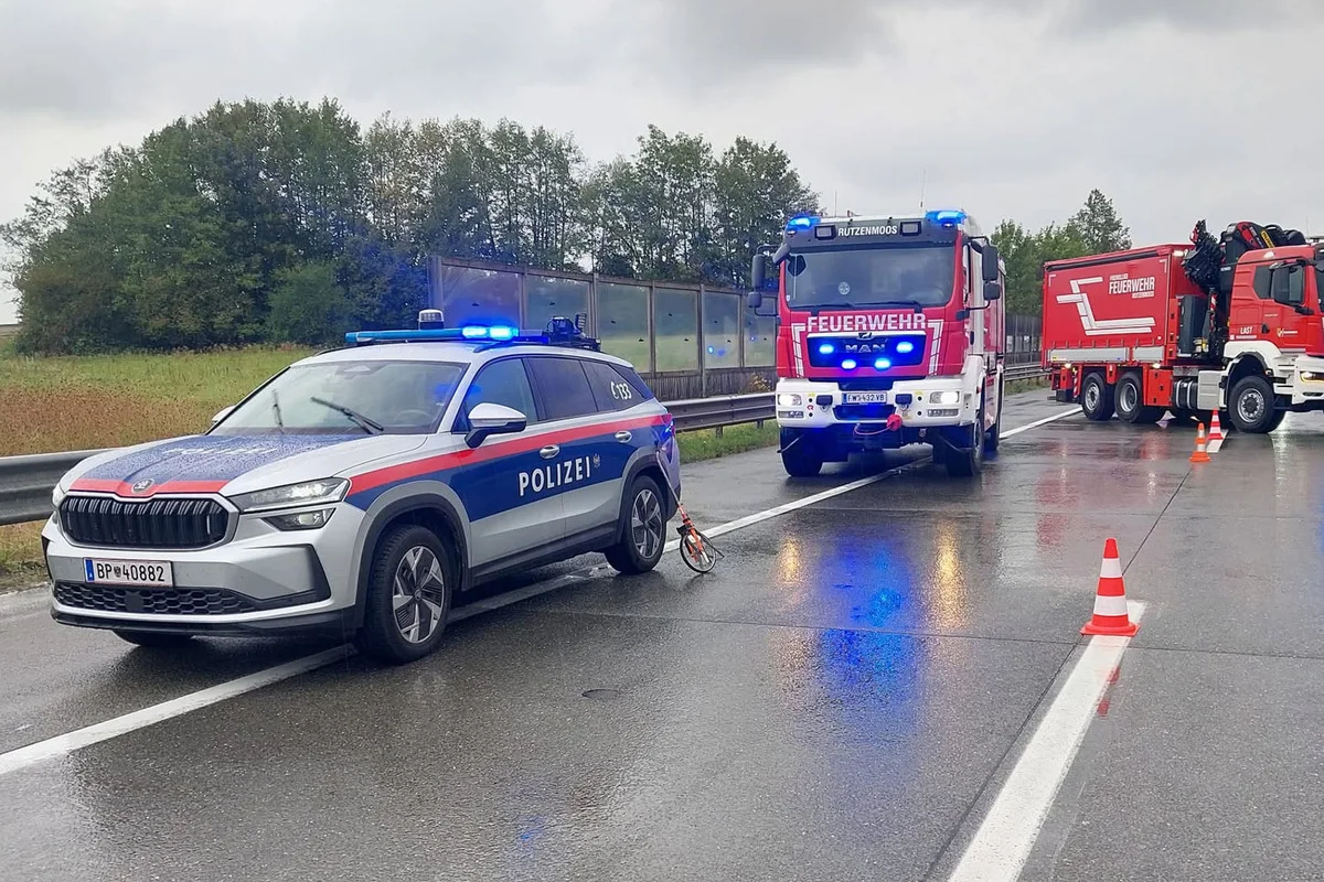 Ein Polizeiwagen und mehrere Feuerwehrfahrzeuge stehen auf einer nassen Straße. Blaulichter sind aktiviert, und Pylonen sperren eine Fahrspur.