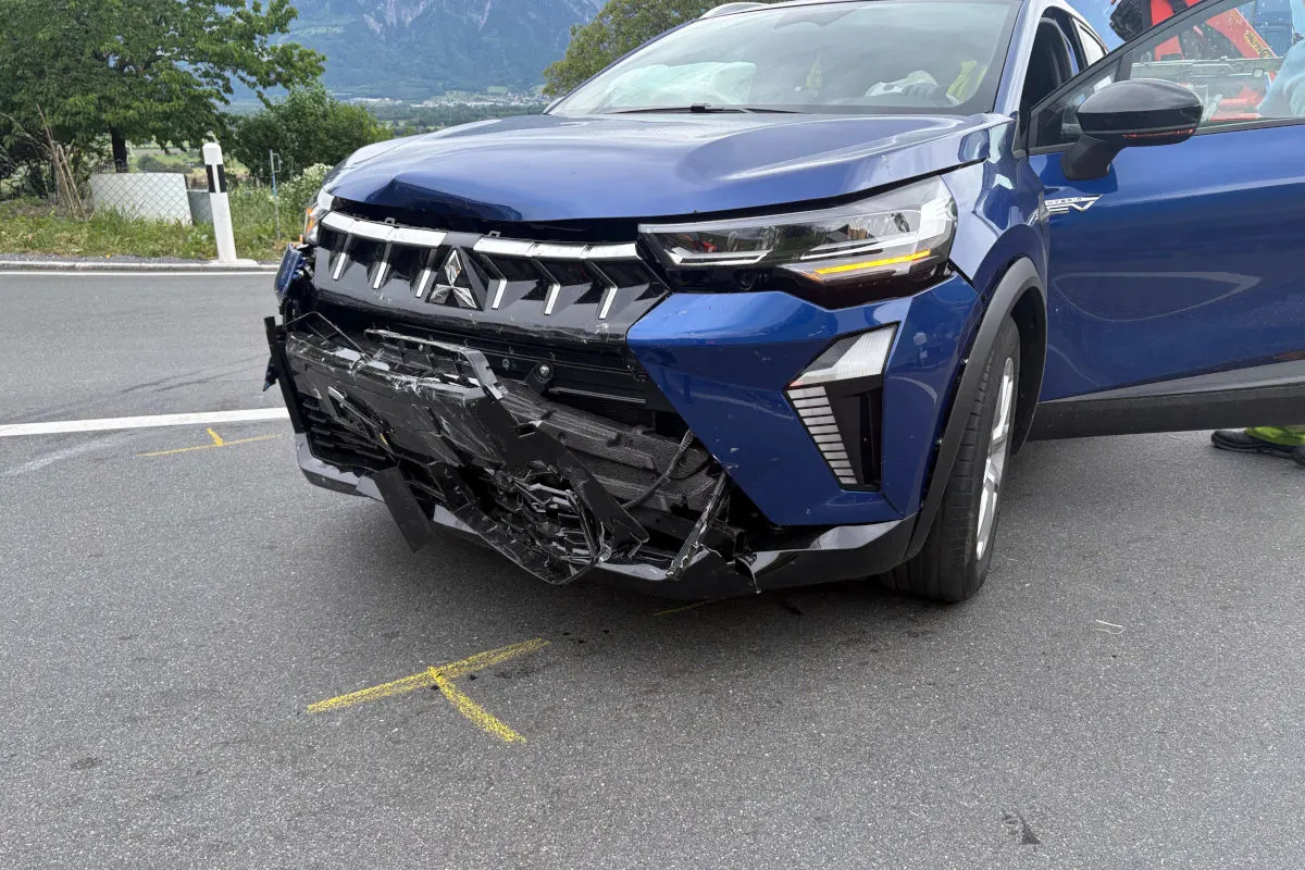The front of a blue car is heavily damaged, parked on the side of a road with surrounding greenery.