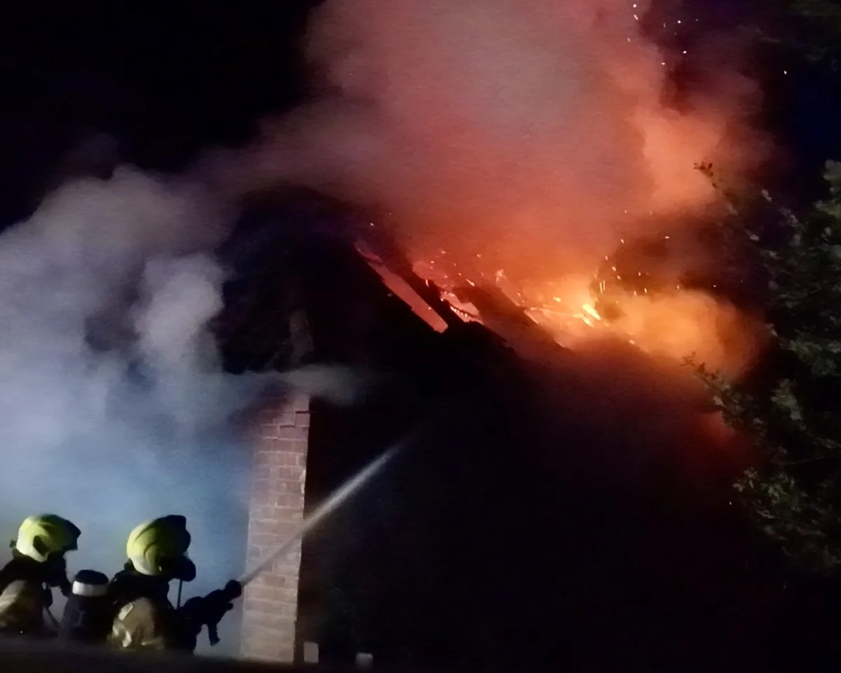 Zwei Feuerwehrleute löschen bei Nacht einen brennenden Dachstuhl mit starken Flammen und Rauchentwicklung.