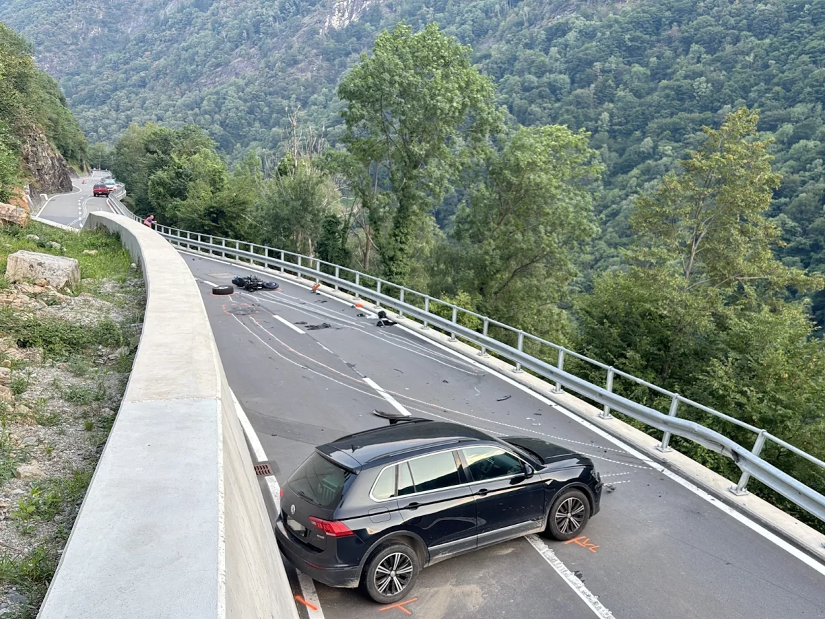 Auf einer Bergstraße ist ein Motorrad in einen Unfall verwickelt, Trümmer und ein Auto sind auf der Fahrbahn verstreut. Die Straße schlängelt sich durch eine grüne Berglandschaft.
