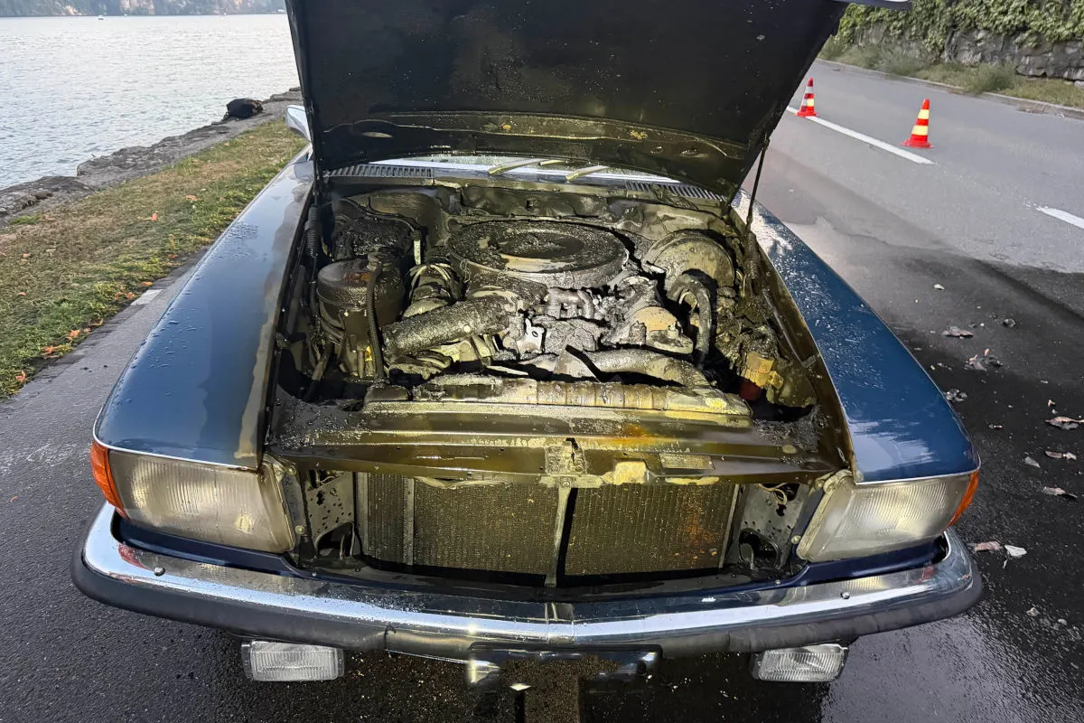 Ein Auto steht am Straßenrand mit geöffneter Motorhaube, der Motor ist stark verbrannt. Im Hintergrund sind Absperrhütchen zu sehen.