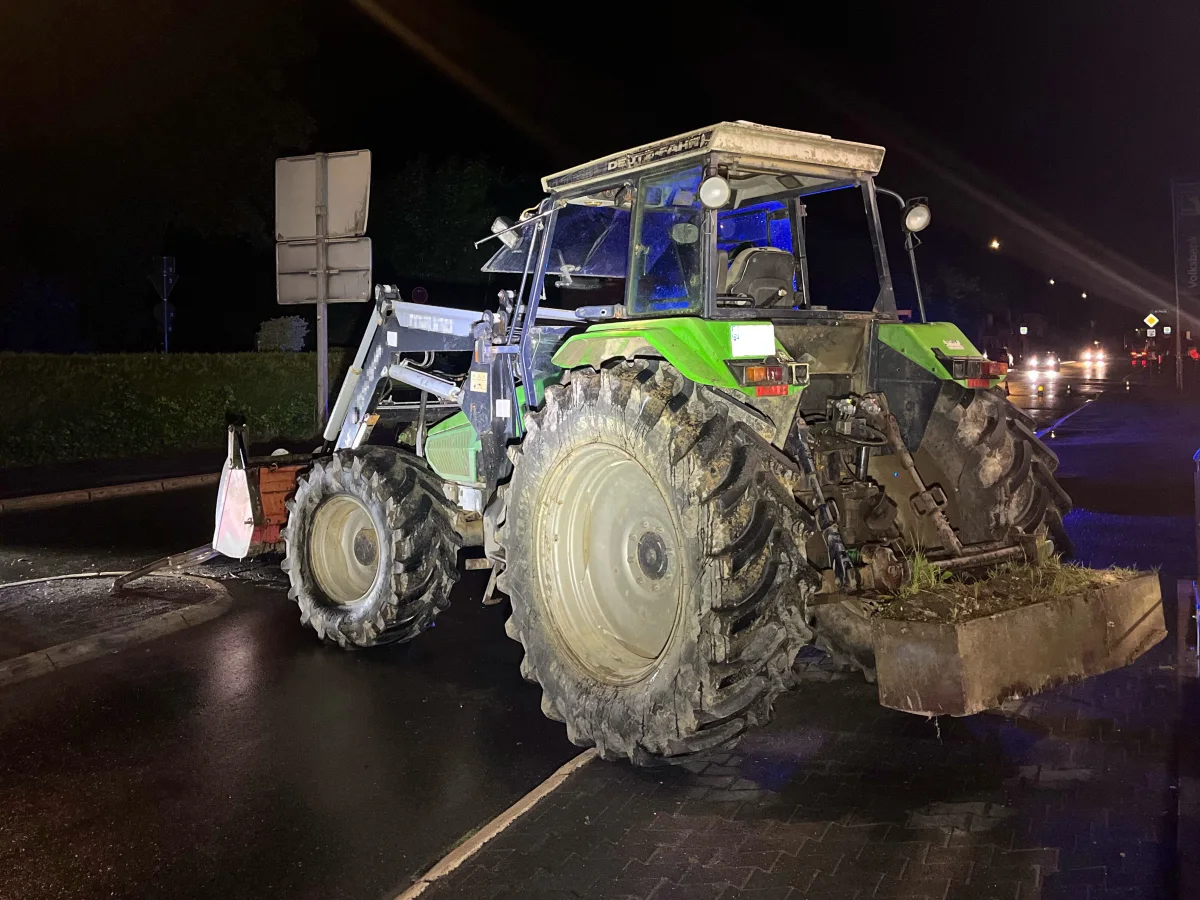 Ein grüner Traktor parkt nachts auf einer belebten, nassen Straße. Straßenbeleuchtung und Fahrzeuge sind im Hintergrund zu sehen.