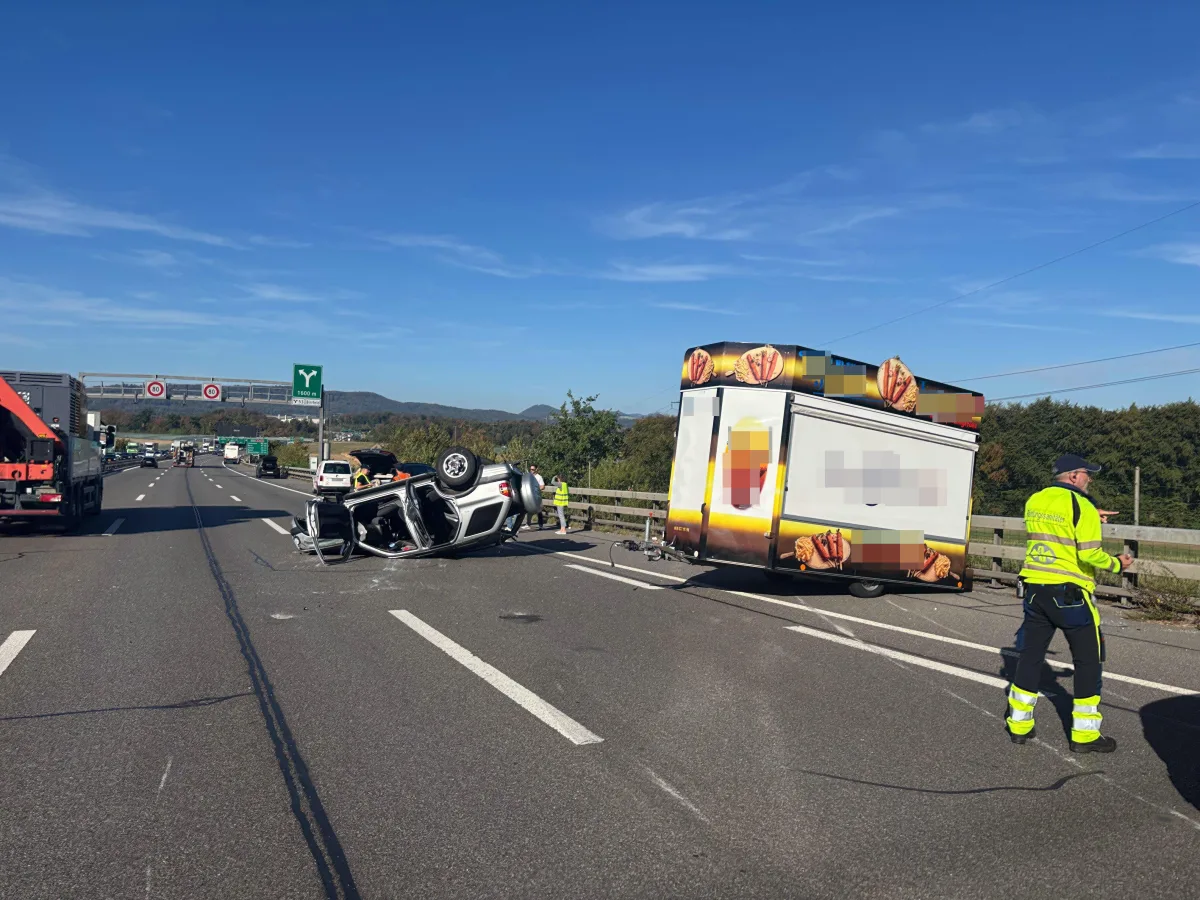 Ein PKW ist auf der Autobahn umgekippt, daneben steht ein beschädigter Verkaufsanhänger. Ein Mann in gelber Warnkleidung ist zu sehen.