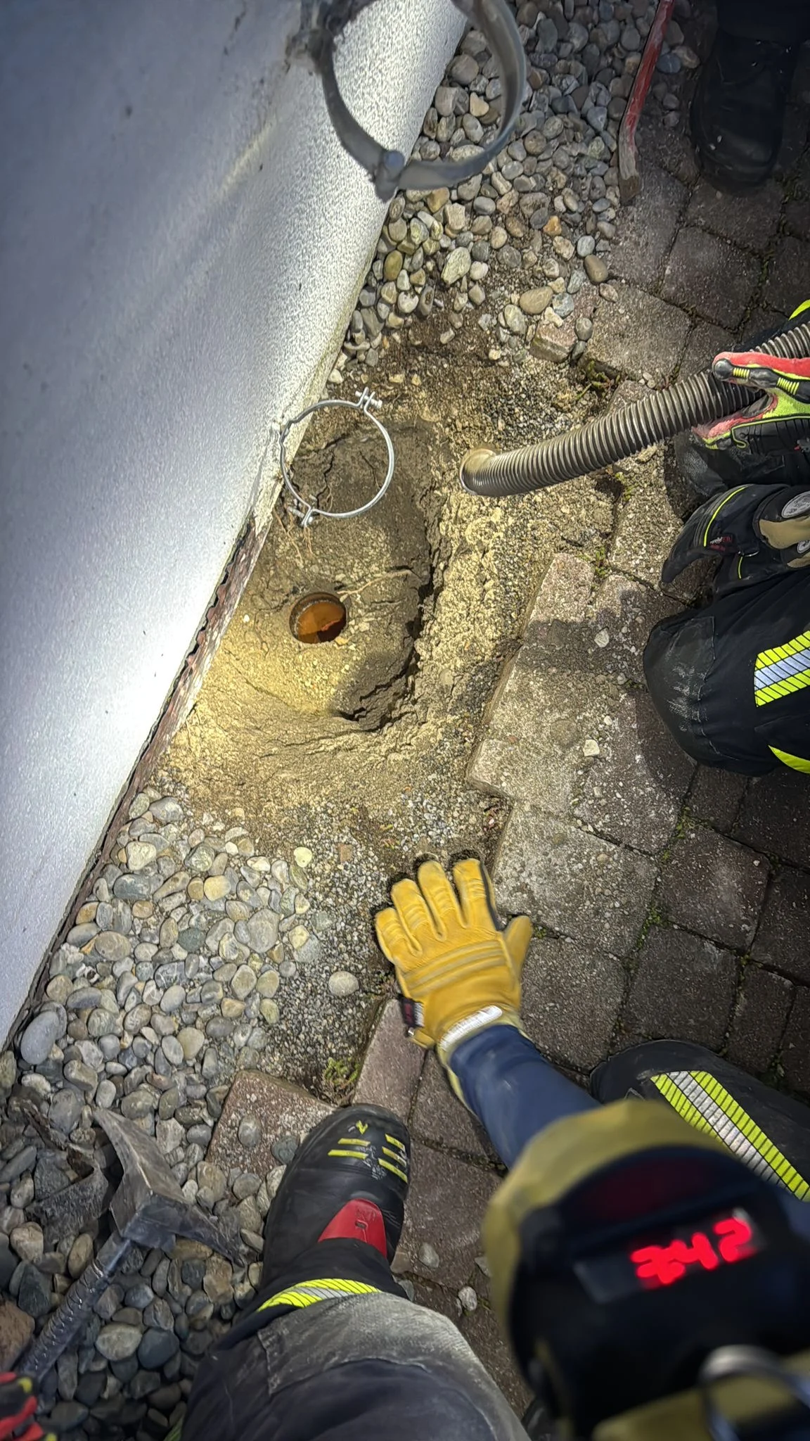 Zwei Arbeiter in Schutzkleidung arbeiten an einem Loch im Boden neben einer weißen Wand. Verschiedene Werkzeuge sind erkennbar.