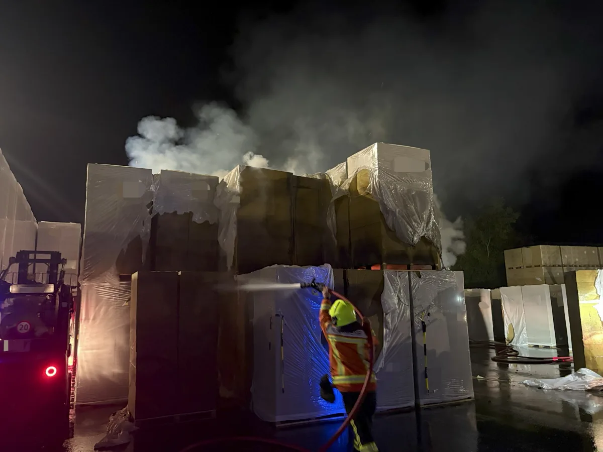 Bei Nacht bekämpft ein Feuerwehrmann ein Feuer, das aus Kartons und Paletten in einer Lagerumgebung lodert.