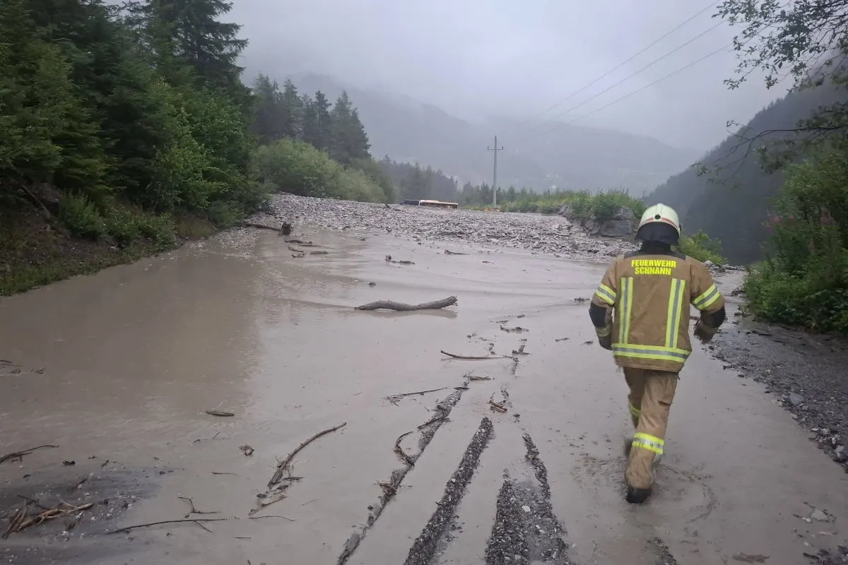 Ein Feuerwehrmann in Schutzkleidung begutachtet eine durch Schlamm und Geröll blockierte Straße. Im Hintergrund sind bewaldete Hügel sichtbar und das Wetter ist neblig und regnerisch.