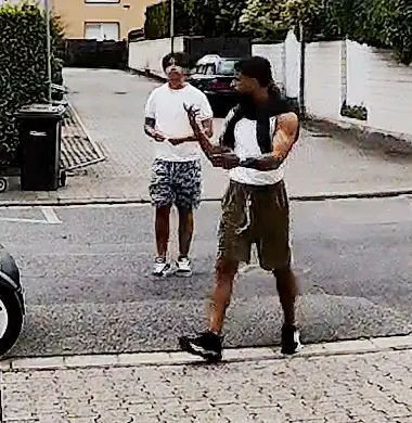 Zwei Männer stehen auf einer gepflasterten Straße in einem Wohngebiet. Einer trägt ein weißes T-Shirt, der andere ein schwarzes.