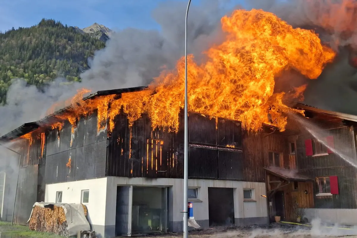 Ein großes, mehrstöckiges Gebäude in ländlicher Umgebung steht in Flammen. Dicker Rauch steigt auf, während das Feuer das Dach erfasst hat.