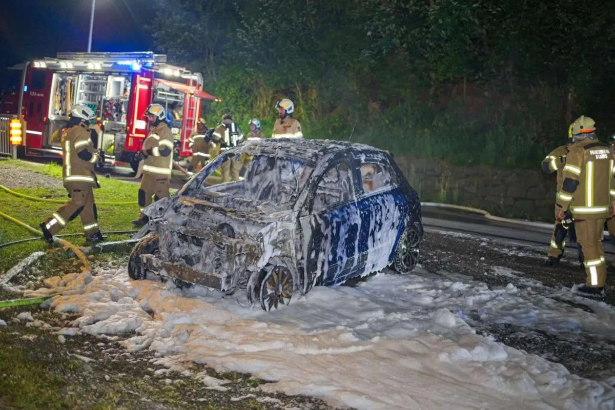 Mehrere Feuerwehrleute stehen bei Nacht um ein ausgebranntes Auto, das mit Löschschaum bedeckt ist. Ein Feuerwehrfahrzeug ist im Hintergrund zu erkennen.