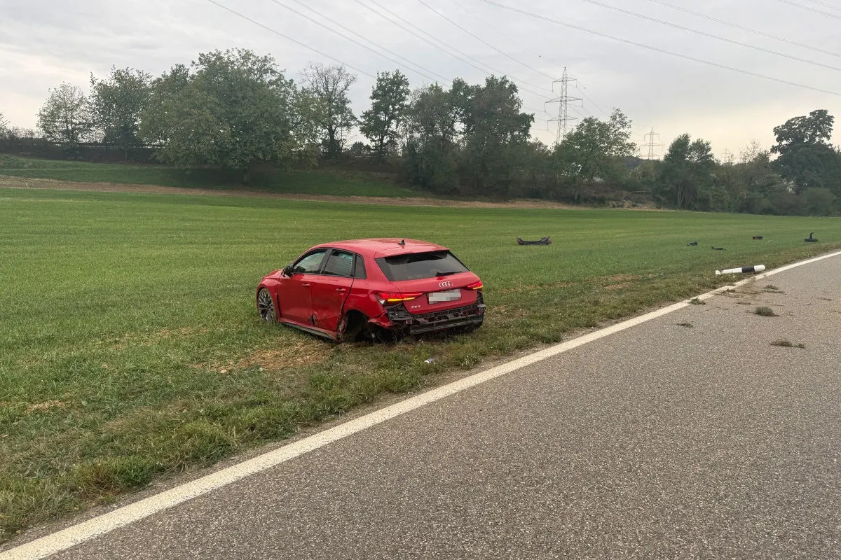 Ein roter Audi mit deutlichem Heckschaden steht auf einem grünen Feld neben einer asphaltierten Straße. Einzelne Fahrzeugteile liegen verstreut auf dem Boden.