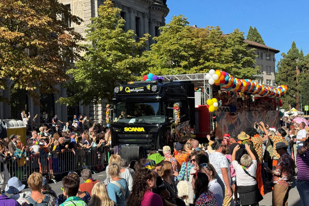 Ein festlich geschmückter Lastwagen fährt bei einer Parade. Viele Menschen säumen die Straße und nehmen Fotos auf.