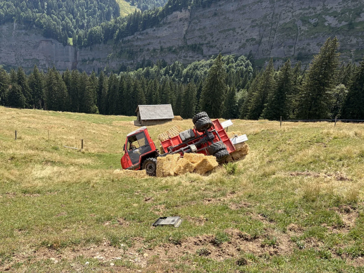 Ein roter Traktor ist auf einer grünen, hügeligen Wiese umgestürzt. Um ihn herum liegen Heuballen, im Hintergrund ist eine Bergwand sichtbar.