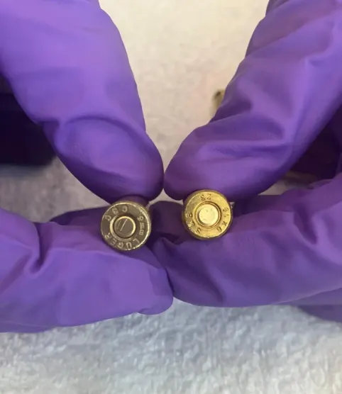Zwei verschiedene Patronenhülsen werden von Händen in lila Handschuhen gehalten, die auf einem hellen Hintergrund liegen.