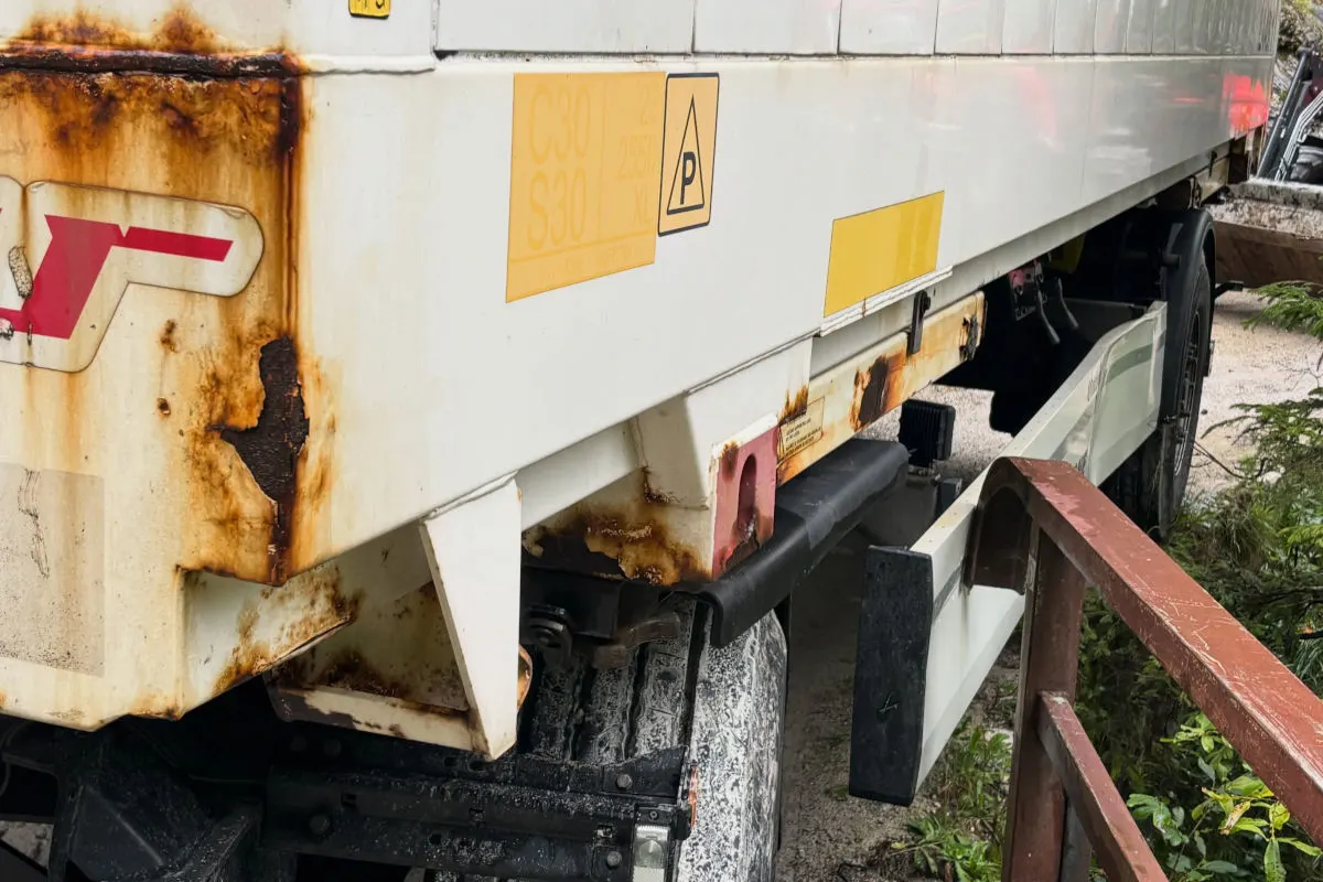 Ein weißer Lkw-Anhänger mit deutlichen Rostspuren steht nahe einer braunen Leitplanke. Im Hintergrund sind Pflanzen zu sehen.