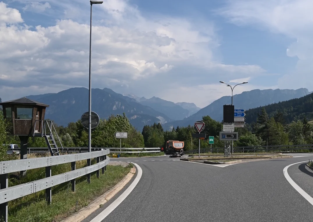 Eine alpine Straßenkreuzung mit einem orangefarbenen Fahrzeug und mehreren Wegweiserschildern vor bergiger Kulisse.