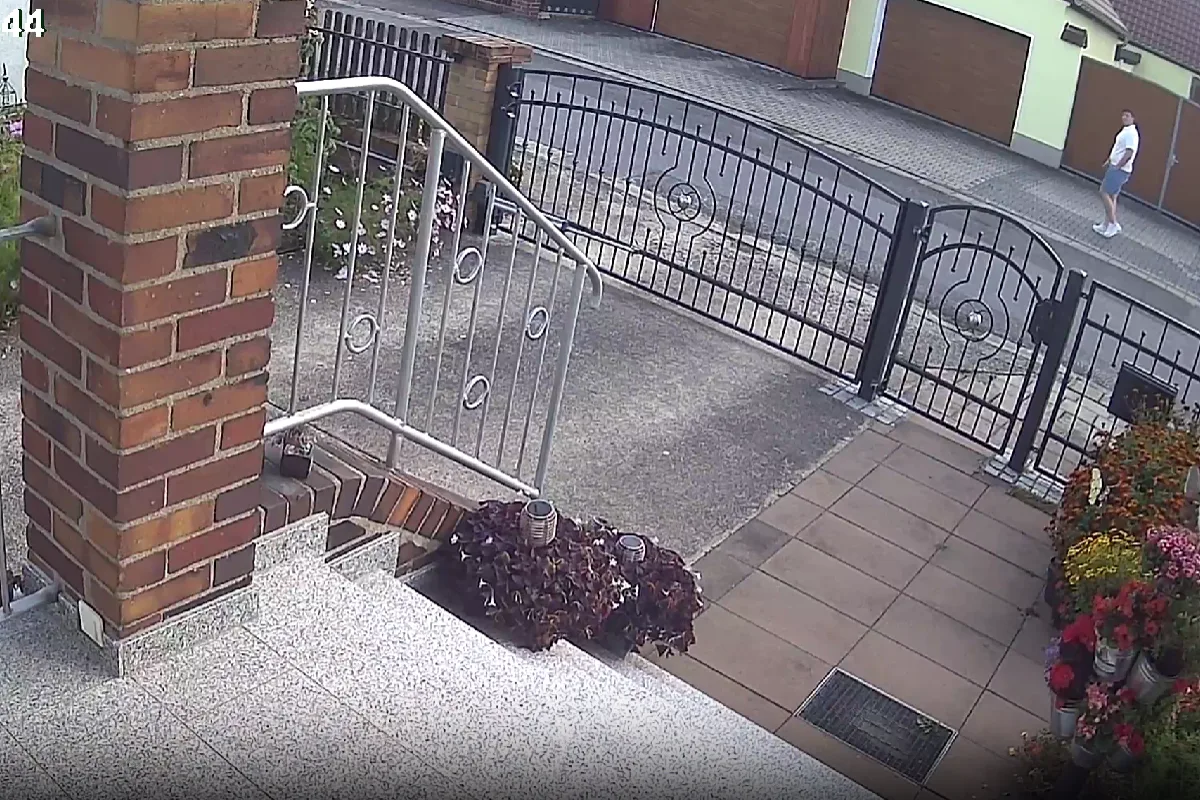 Eine Person in Shorts und T-Shirt geht auf einem Gehweg vor einem Haus mit Metallzaun. Im Vordergrund sind Blumenbeete und Treppen zu sehen.