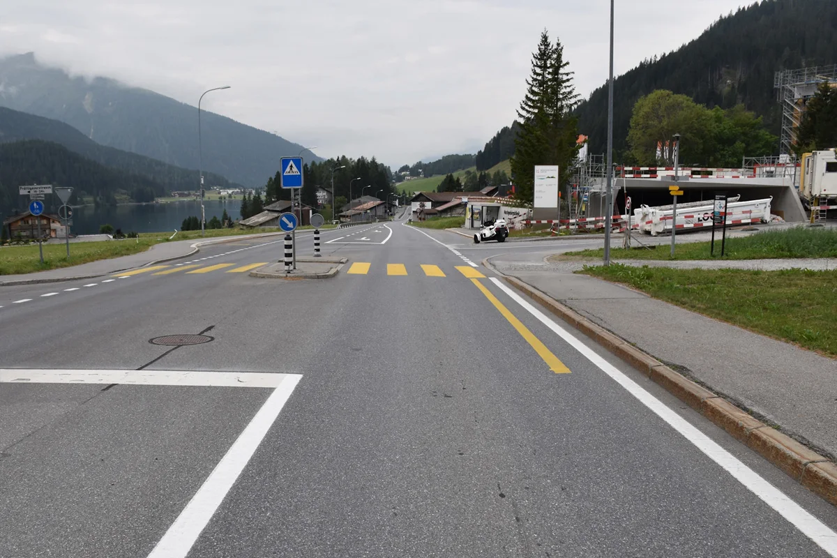 Eine Dorfstraße mit gelben Zebrastreifen führt zu einem Berg mit einem umgekippten Fahrzeug am rechten Rand. Im Hintergrund sind Berge und ein bewölkter Himmel sichtbar.