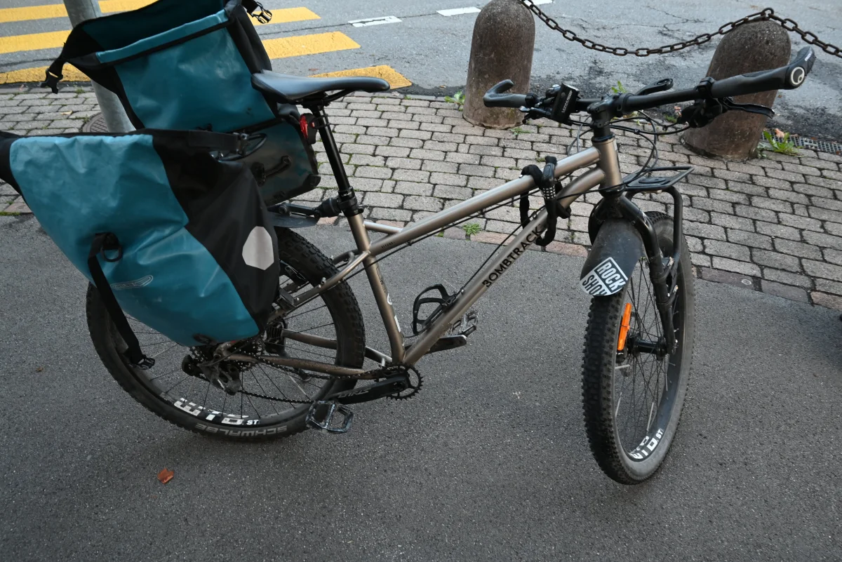 Ein Fahrrad mit blauen Packtaschen ist auf einem Bürgersteig in einer städtischen Umgebung abgestellt. Im Hintergrund sind Straßenpflaster und gelbe Zebrastreifen zu sehen.