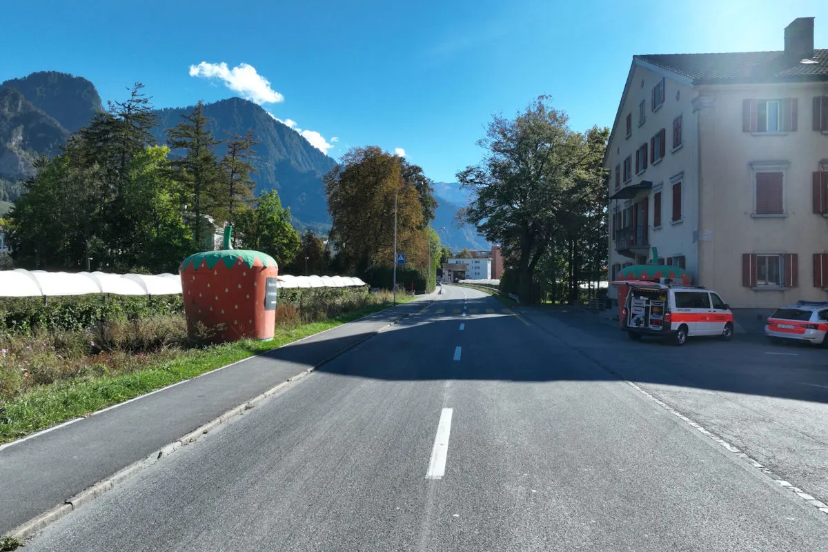 Ein Rettungsfahrzeug parkt auf einem Bürgersteig neben einem großen Wohngebäude im Vordergrund. Im Hintergrund sind Berge und blauer Himmel zu sehen.